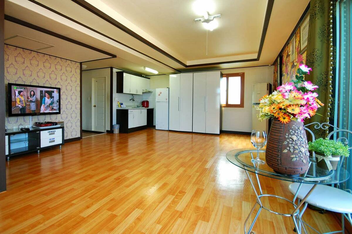 Un salón amplio con piso de madera, una mesa de vidrio con sillas, una cocina al fondo y un jarrón decorativo con flores sobre la mesa.