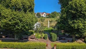 A garden pathway leading to a house with a red roof, surrounded by lush greenery and a clear blue sky.