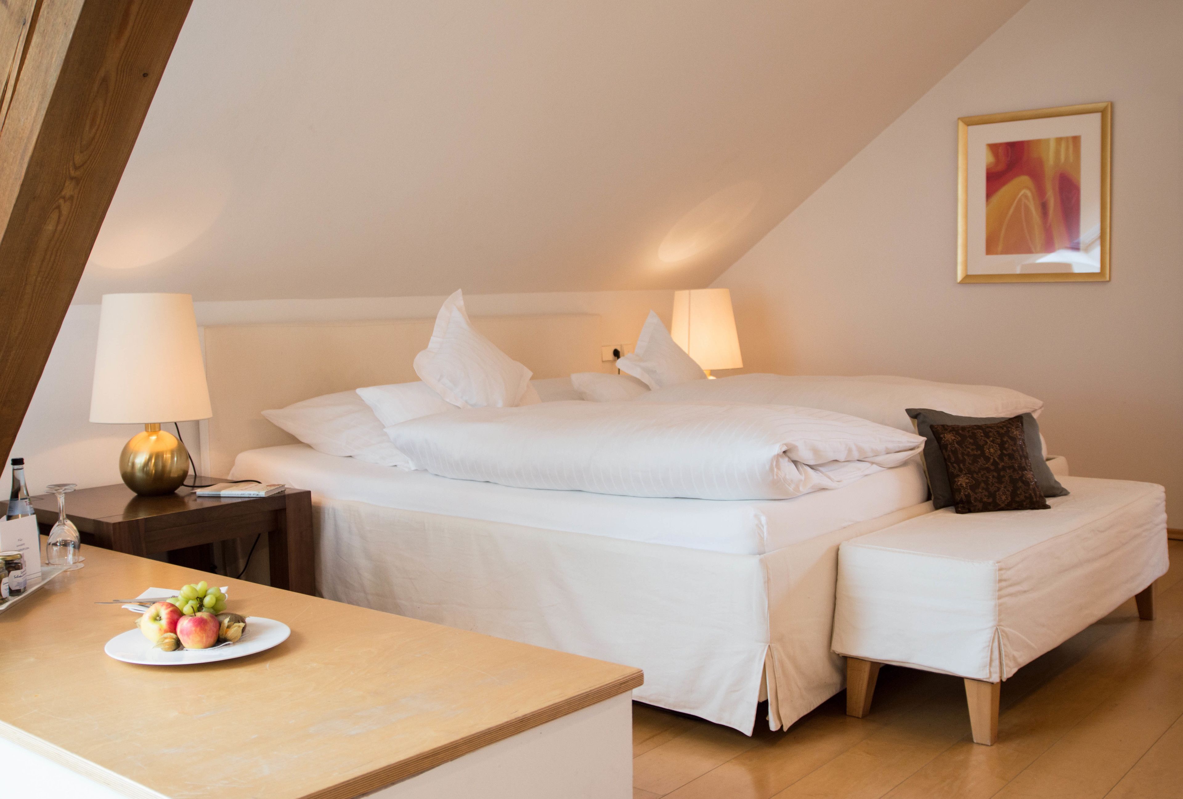 A hotel room with a bed, bedside tables, a bedside lamp, a small table with fruit, and a painting on the wall.