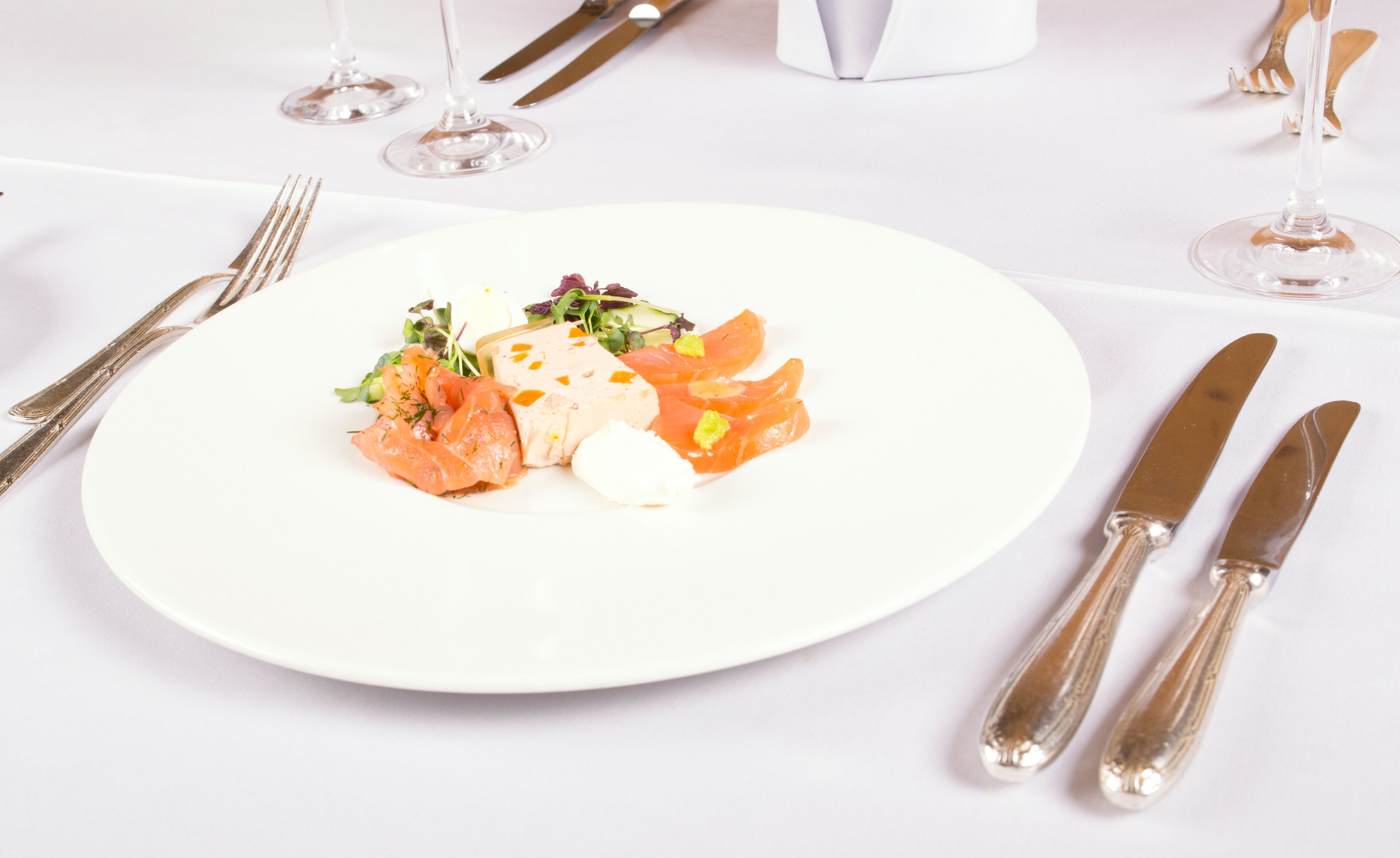 A white plate with a salad, salmon, and cheese on a table with silverware and wine glasses.