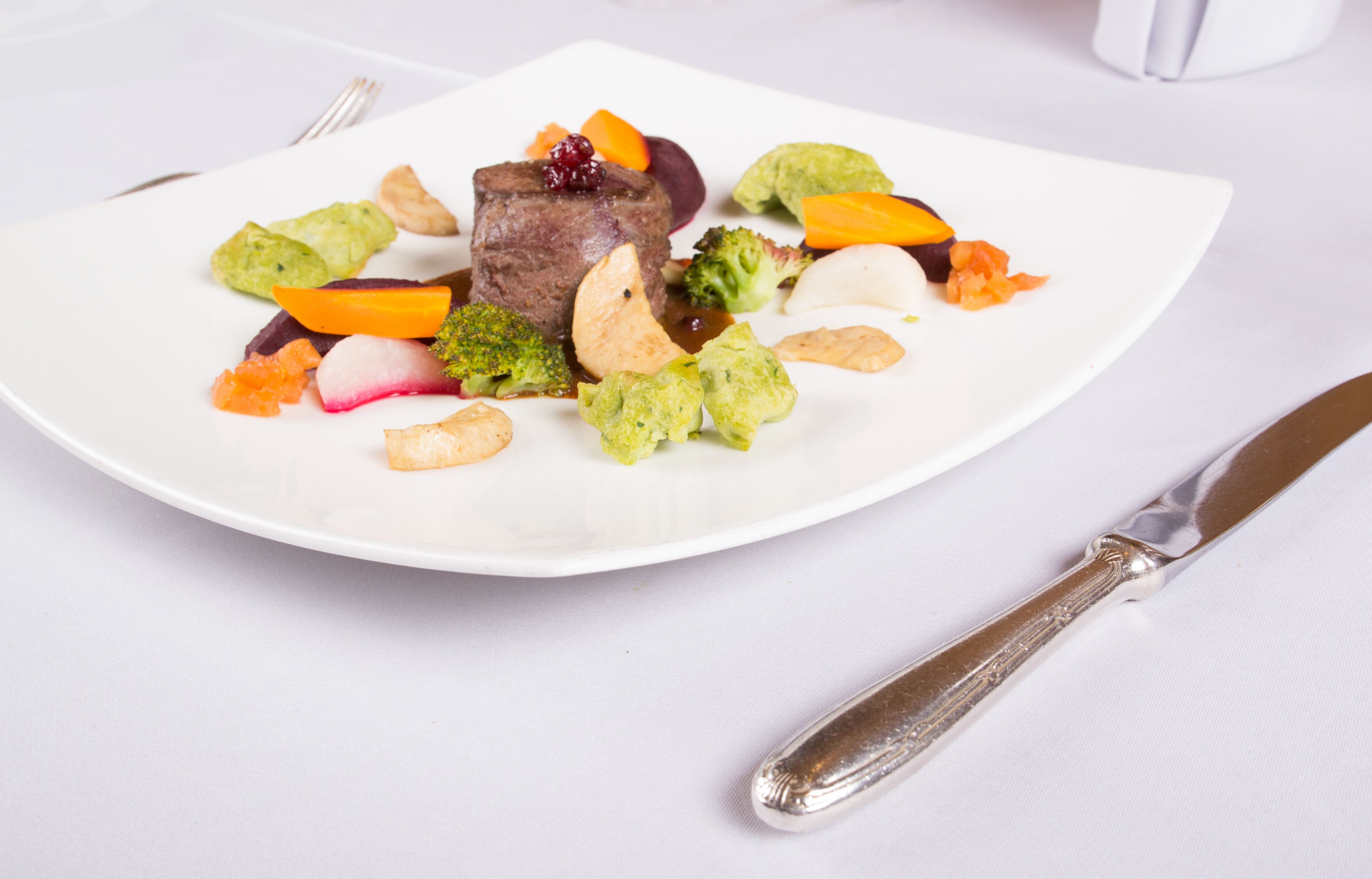 A white plate with a piece of meat, vegetables, and nuts.
