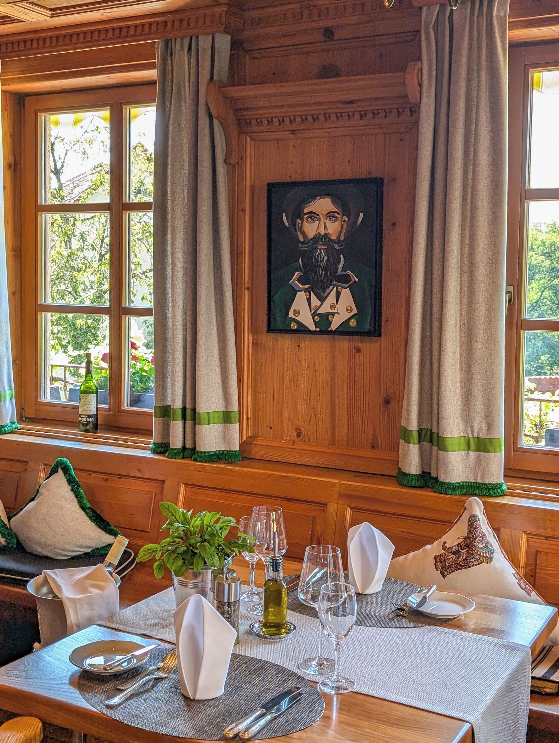 A dining area with a wooden table set for two, a painting on the wall, and curtains with green trim.