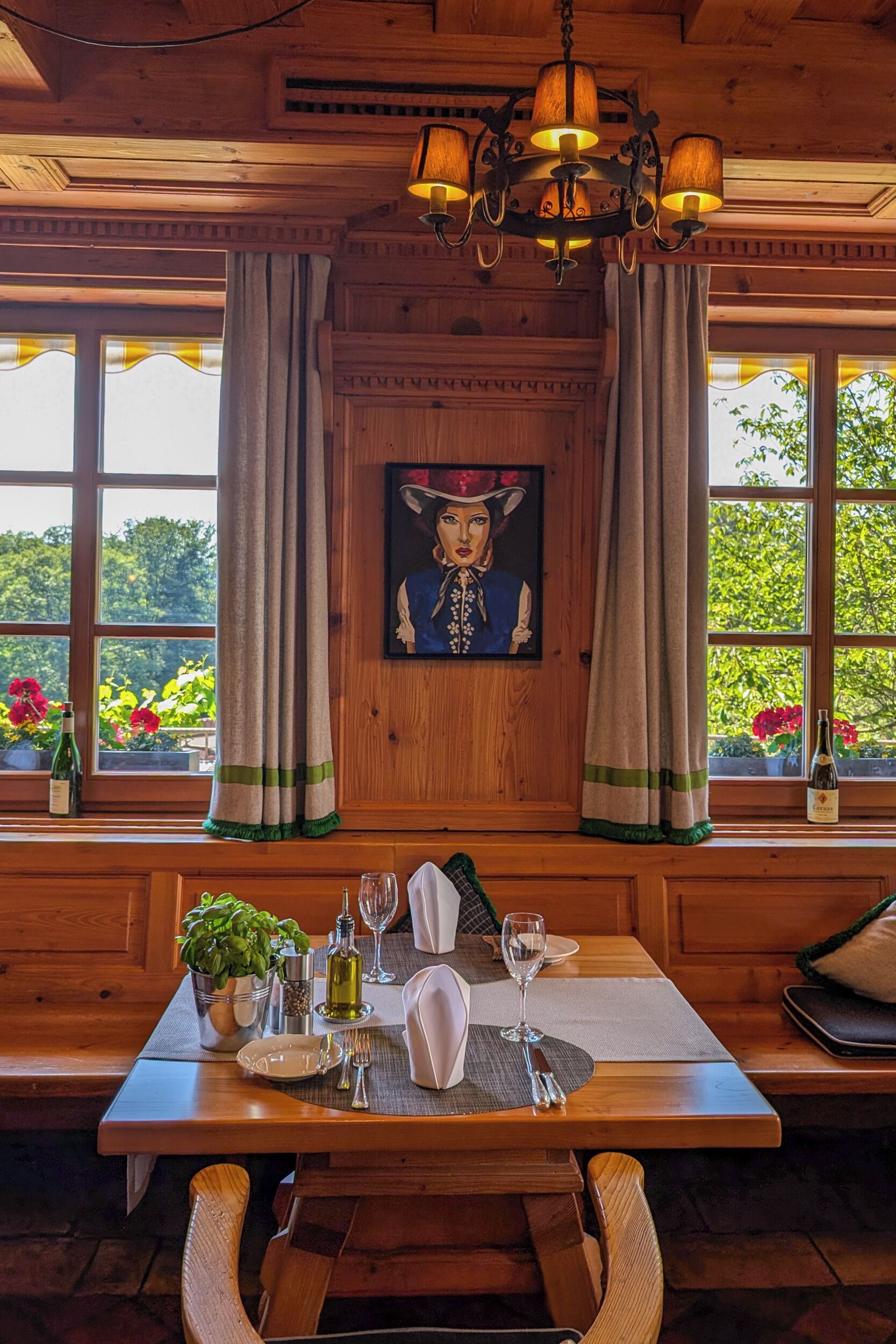 A wooden dining table set with plates, glasses, and a bottle, with a painting and curtains on the wall, and a window with flowers outside.