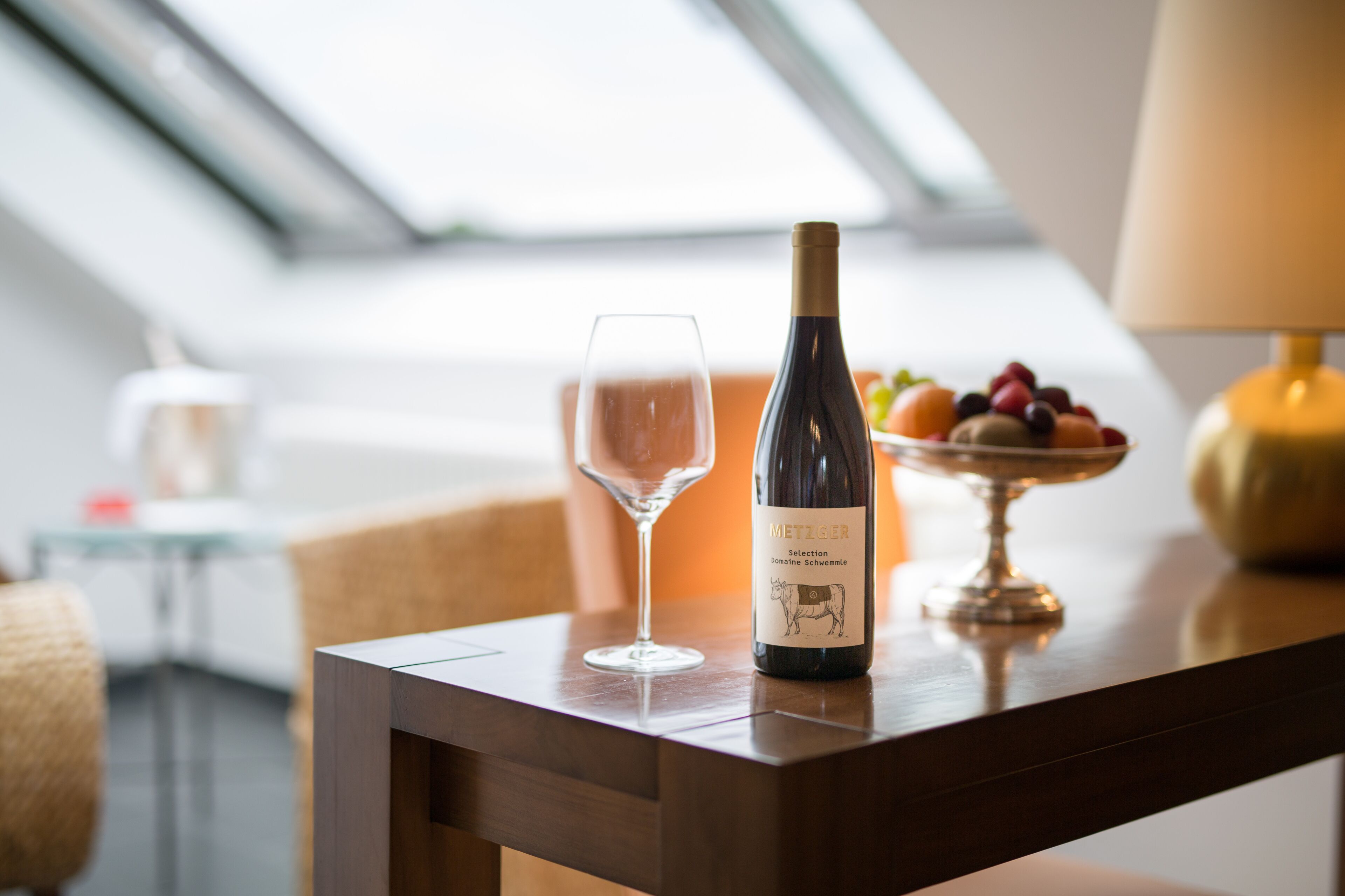 A bottle of Metzger wine, a glass, and a bowl of fruit on a wooden table in a modern room with a skylight.
