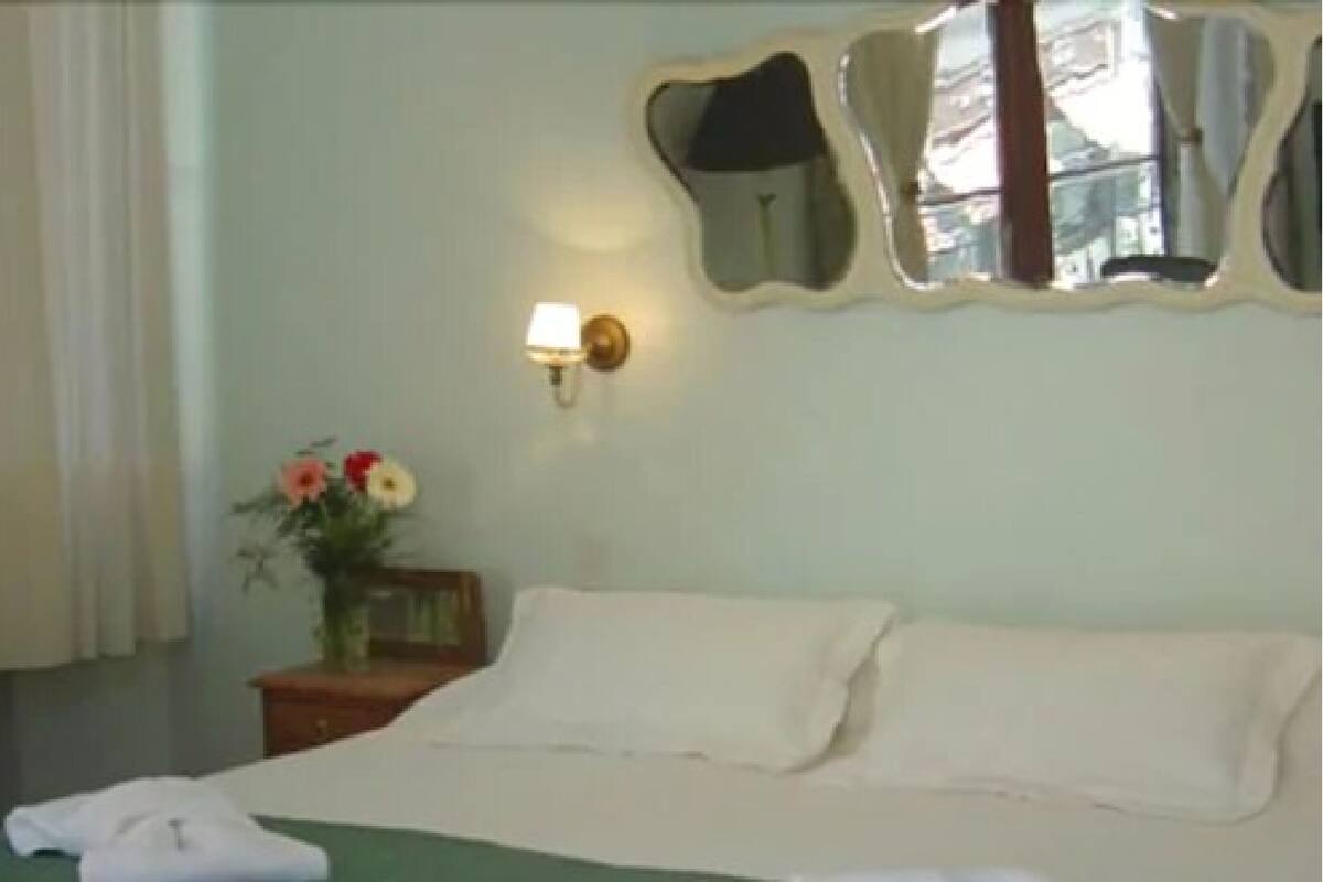 Ein Hotelzimmer mit einem Bett, einem Nachttisch mit Lampe, einem Spiegel und einer Vase mit Blumen.