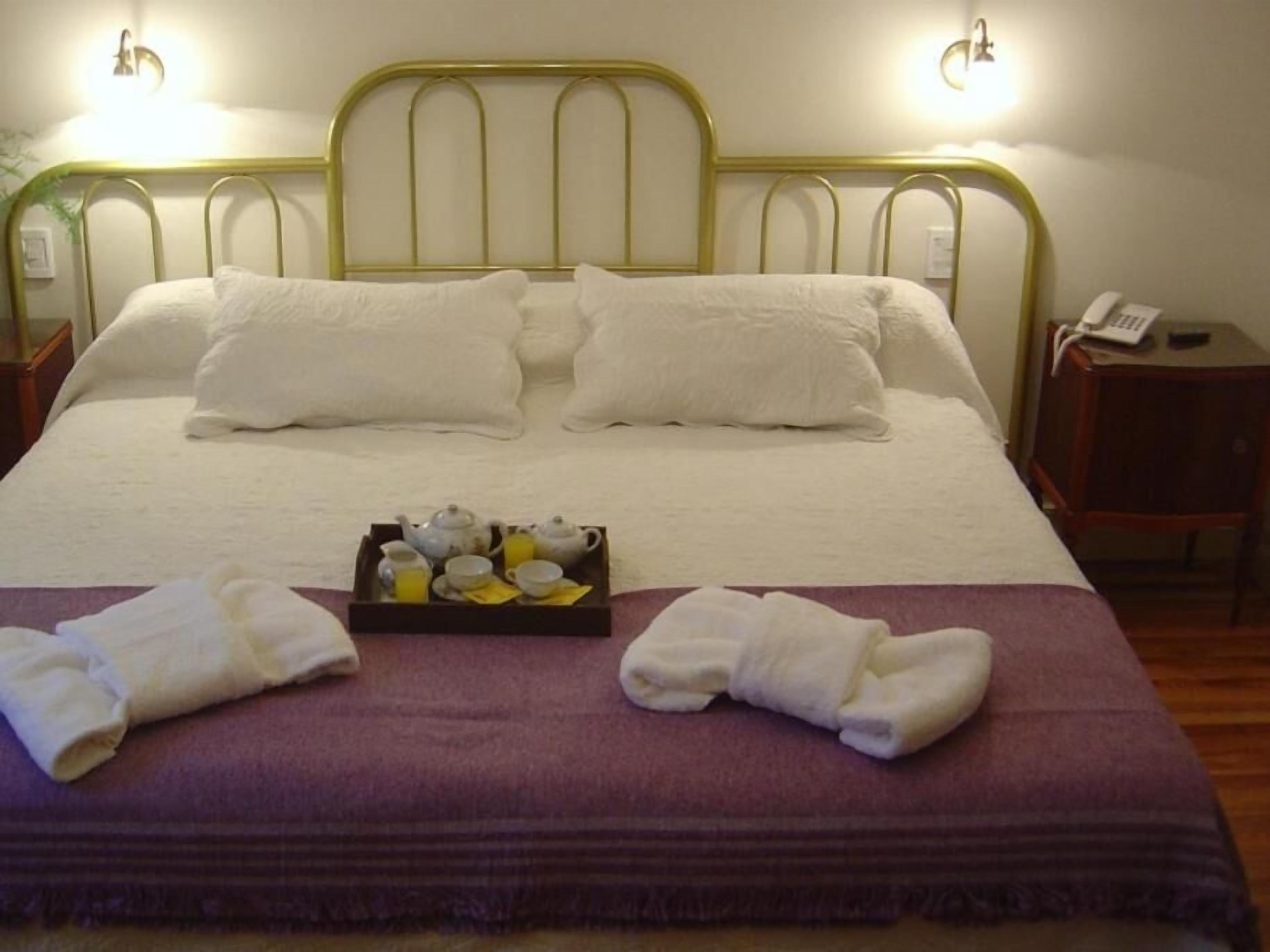 A neatly made bed with white linens and pillows, a tray with tea and juice, and a purple blanket.