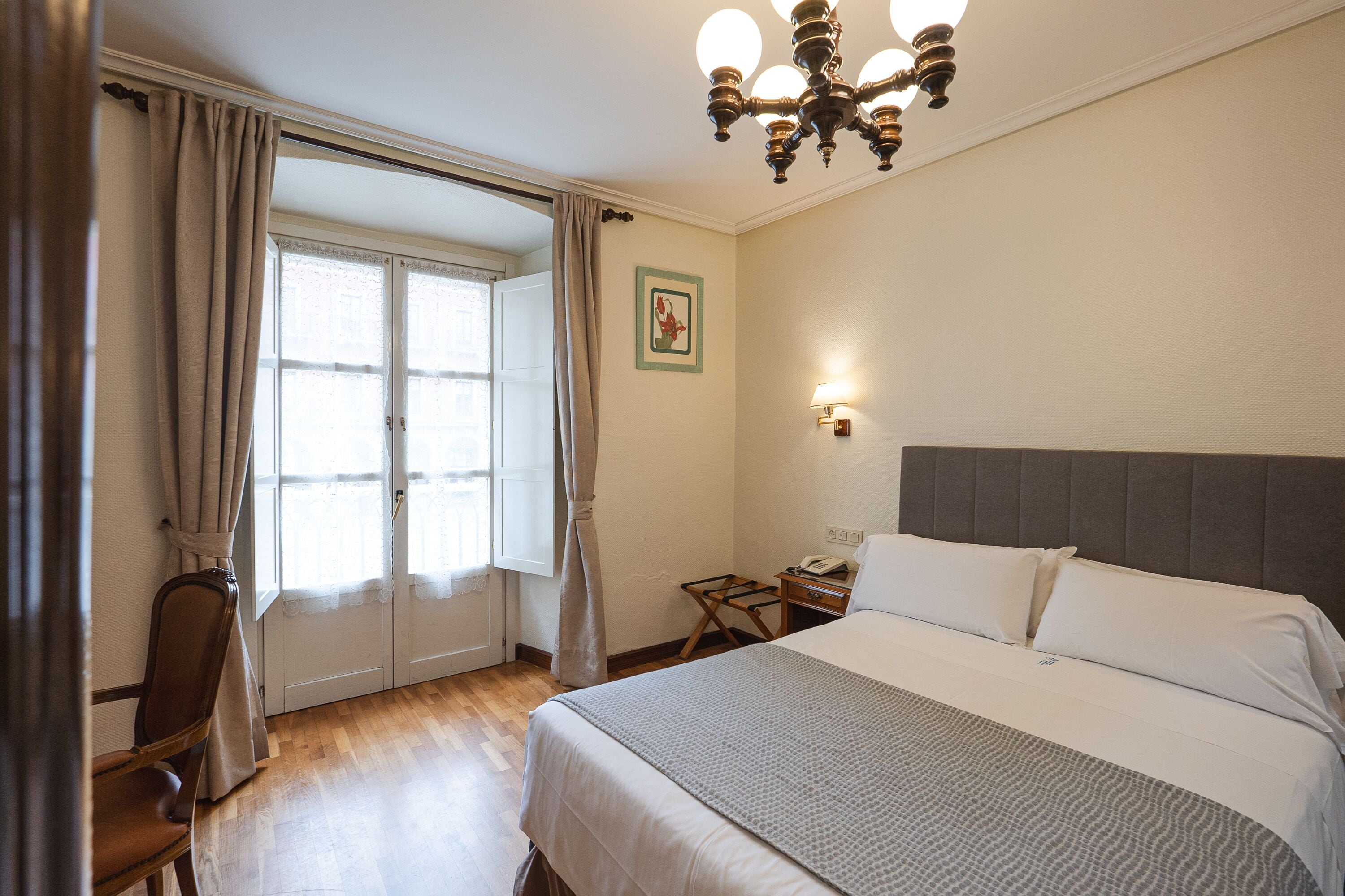 A hotel room with a large bed, a wooden chair, a desk with a lamp, and a window with curtains.