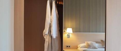A hotel room with a wooden floor, a bed with white bedding, a nightstand with a lamp, and a closet with hanging clothes.