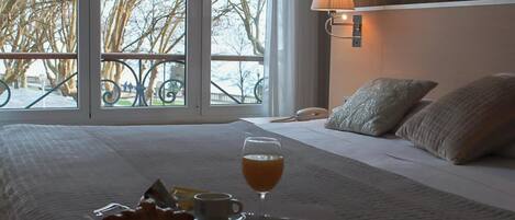 A hotel room with a bed, breakfast tray, and a view of trees through large windows.