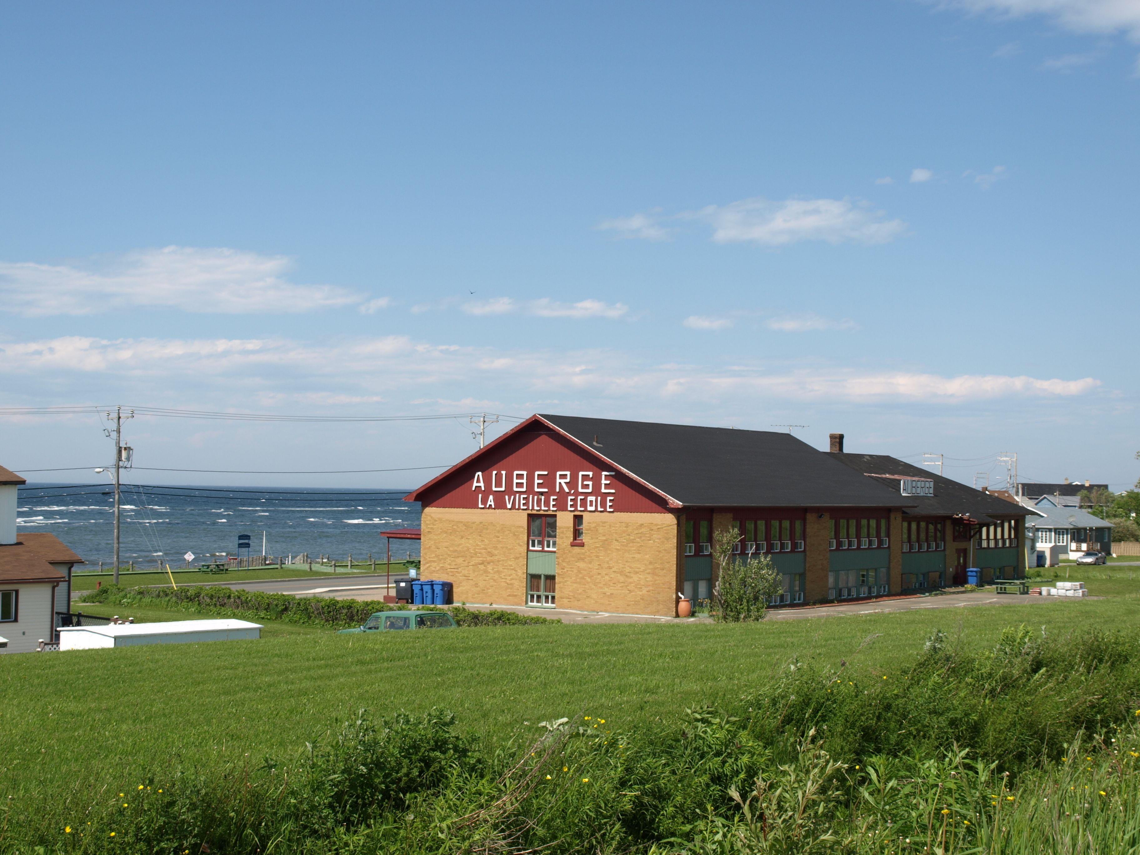 En bygning kaldet "Auberge La Vieille Ecole" med en murstensfacade og et rødt tag, beliggende i nærheden af et vandareal.