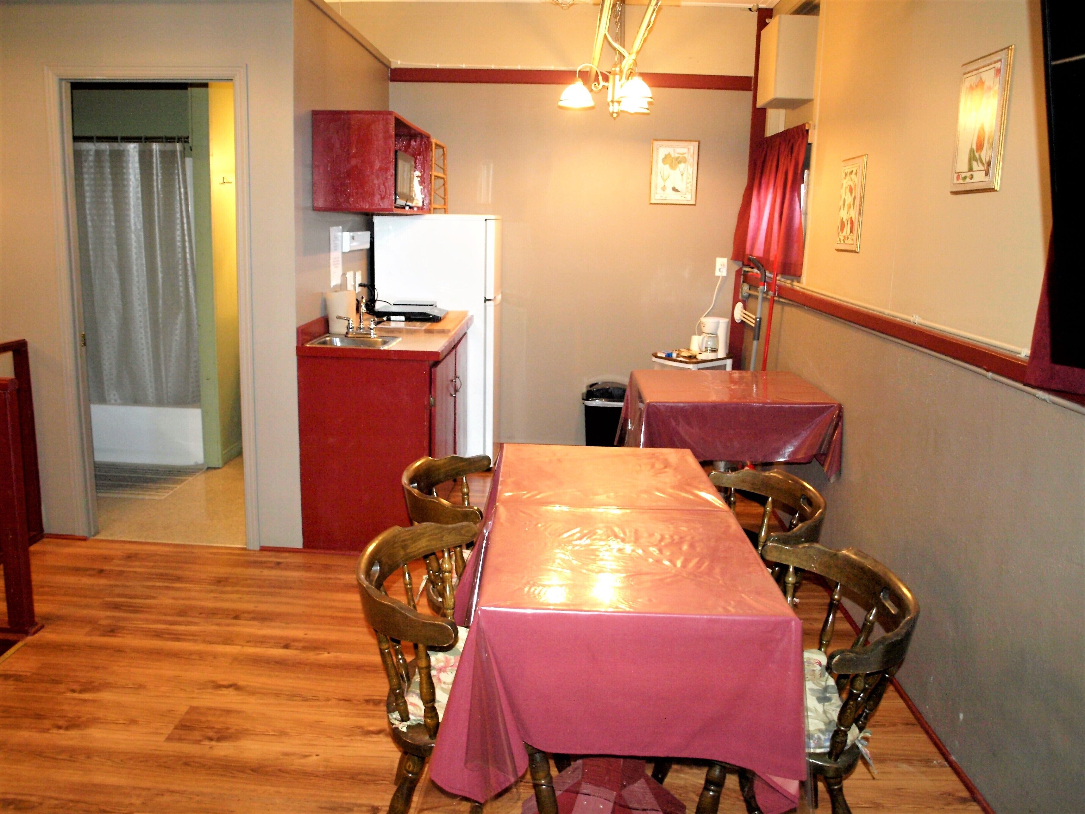 A compact kitchen with a refrigerator, sink, and wooden cabinets. There is a dining area with a table covered in a plastic tablecloth and four wooden chairs.