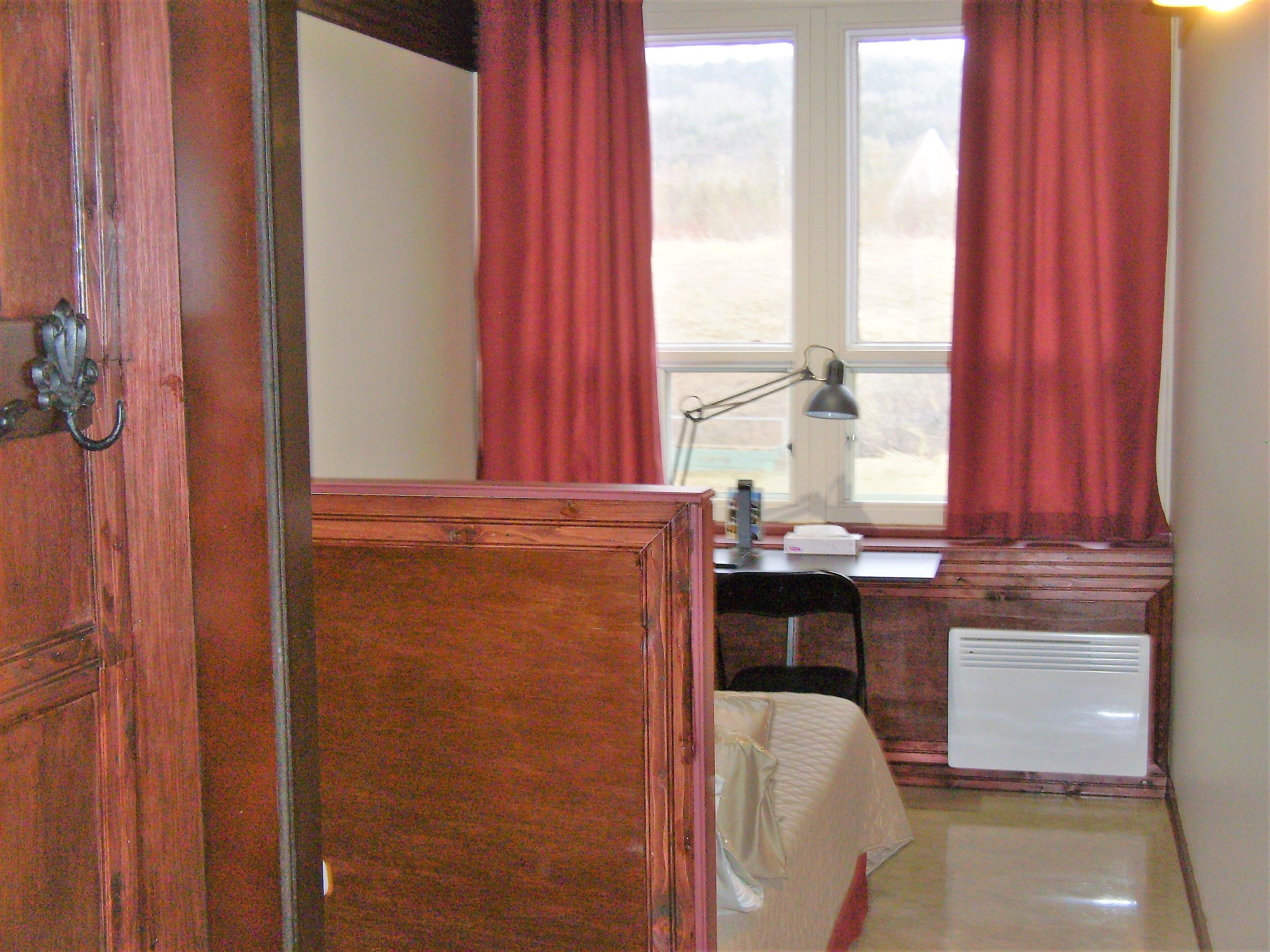 Una habitación con un escritorio de madera, una silla, un radiador y un ventanal con cortinas rojas.