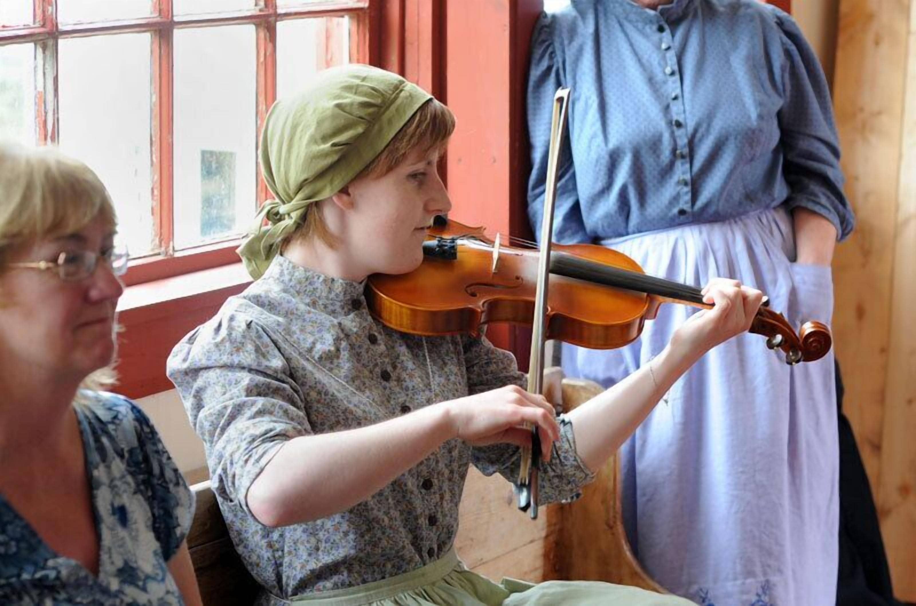 A person playing a violin inside a room with wooden walls and a window.