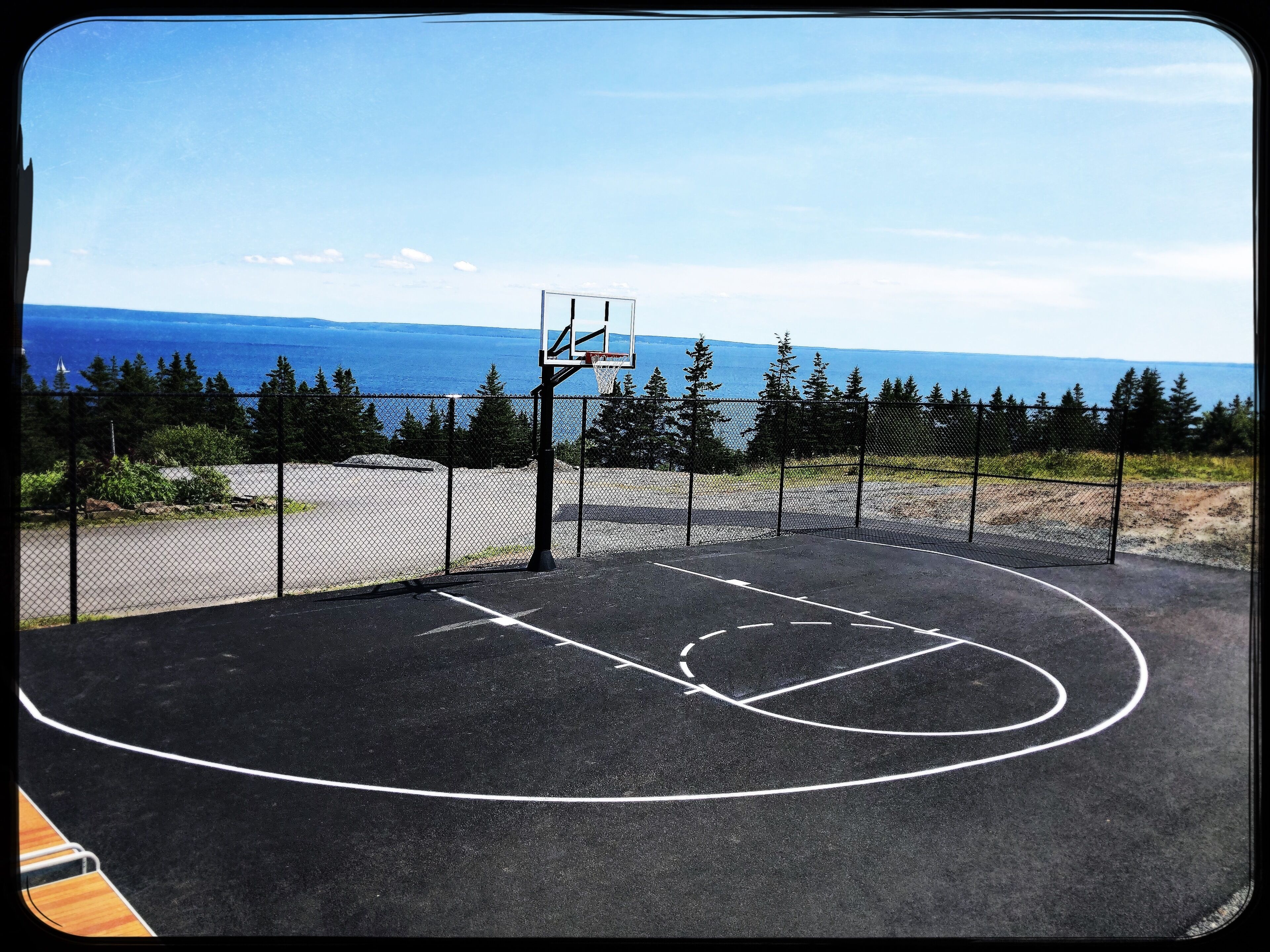 En udendørs basketballbane med en basketballkurv, omkranset af et wirehegn, med udsigt til havet og træer i baggrunden.