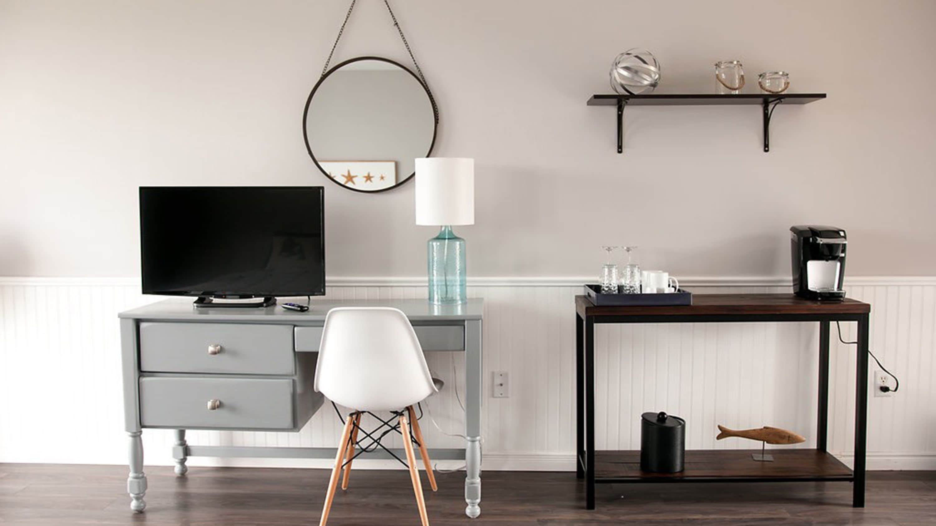 A modern hotel room with a grey desk, a television, a lamp, a coffee maker, and a small shelf with decorative items.