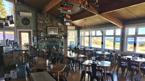 Ein Restaurant mit einer Steinwand, Holztischen und -stühlen. An der Wand hängen Schilder und es gibt einen Blick ins Freie durch große Fenster.