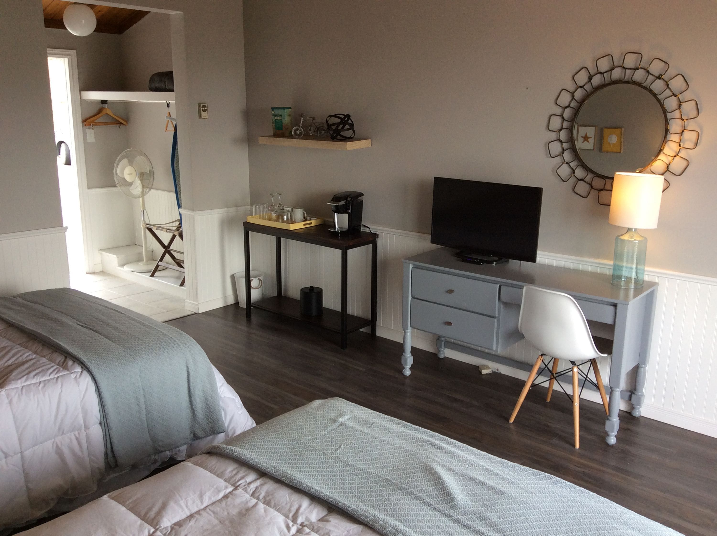 A hotel room with two beds, a desk with a TV, a chair, a small table with items, a mirror, and a lamp.