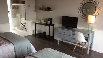 A hotel room with two beds, a desk with a TV, a chair, a small table with items, a mirror, and a lamp.