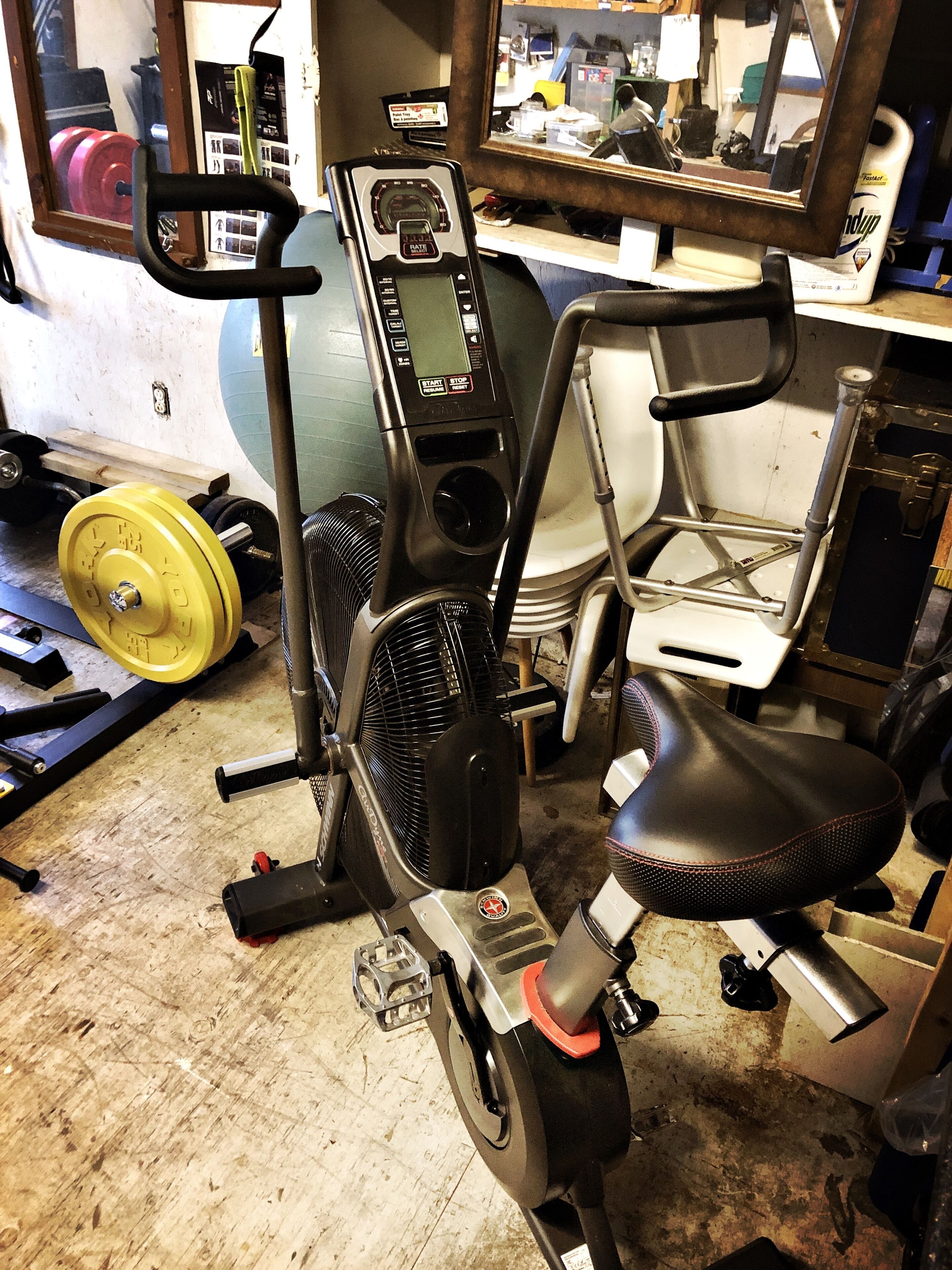 Una bicicleta estática con pantalla digital, un ventilador y una apiladora de pesas en una habitación con espejo y equipo de gimnasio.