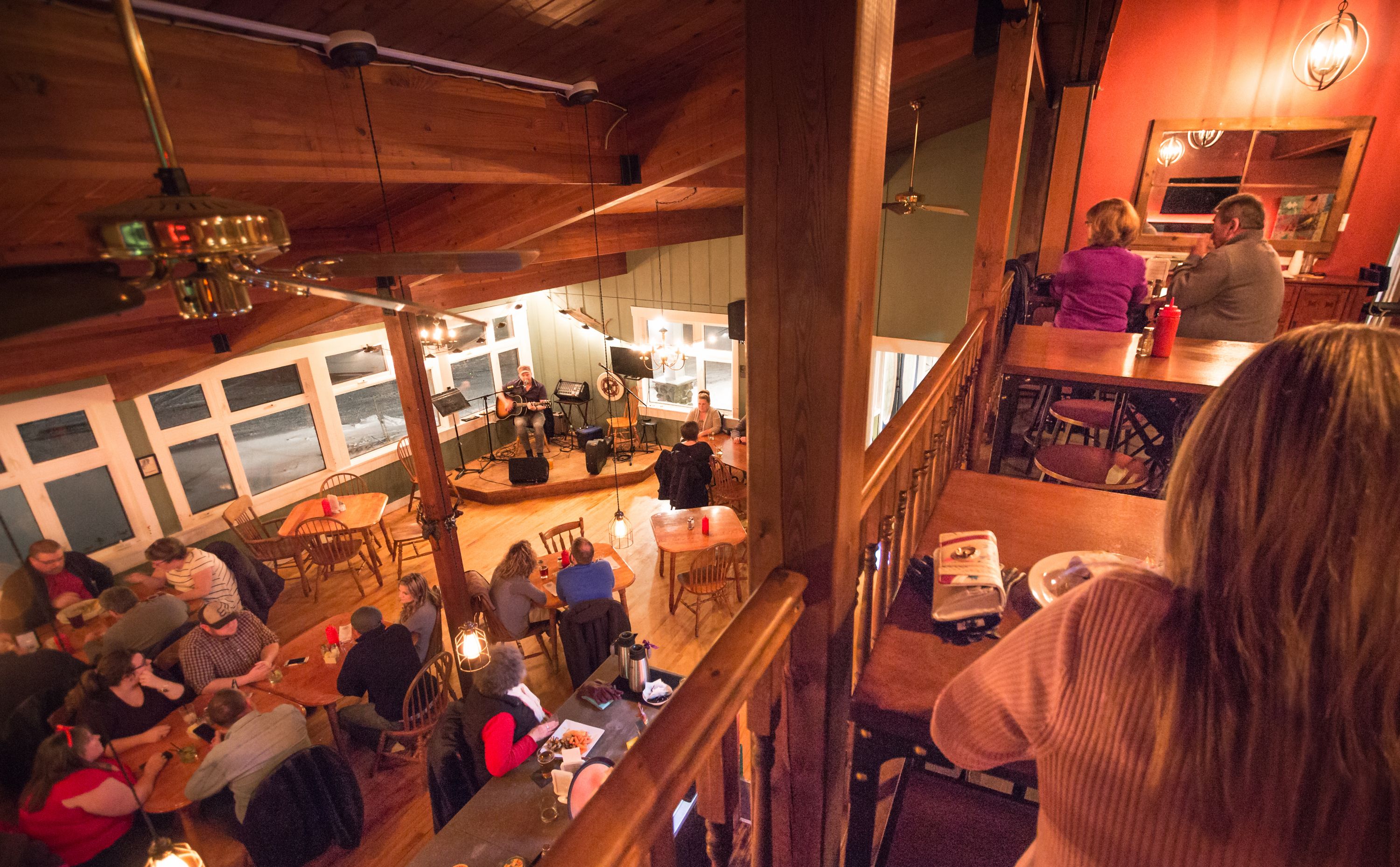 Un restaurante acogedor con interiores de madera, un escenario para música en vivo y comensales cenando en mesas.