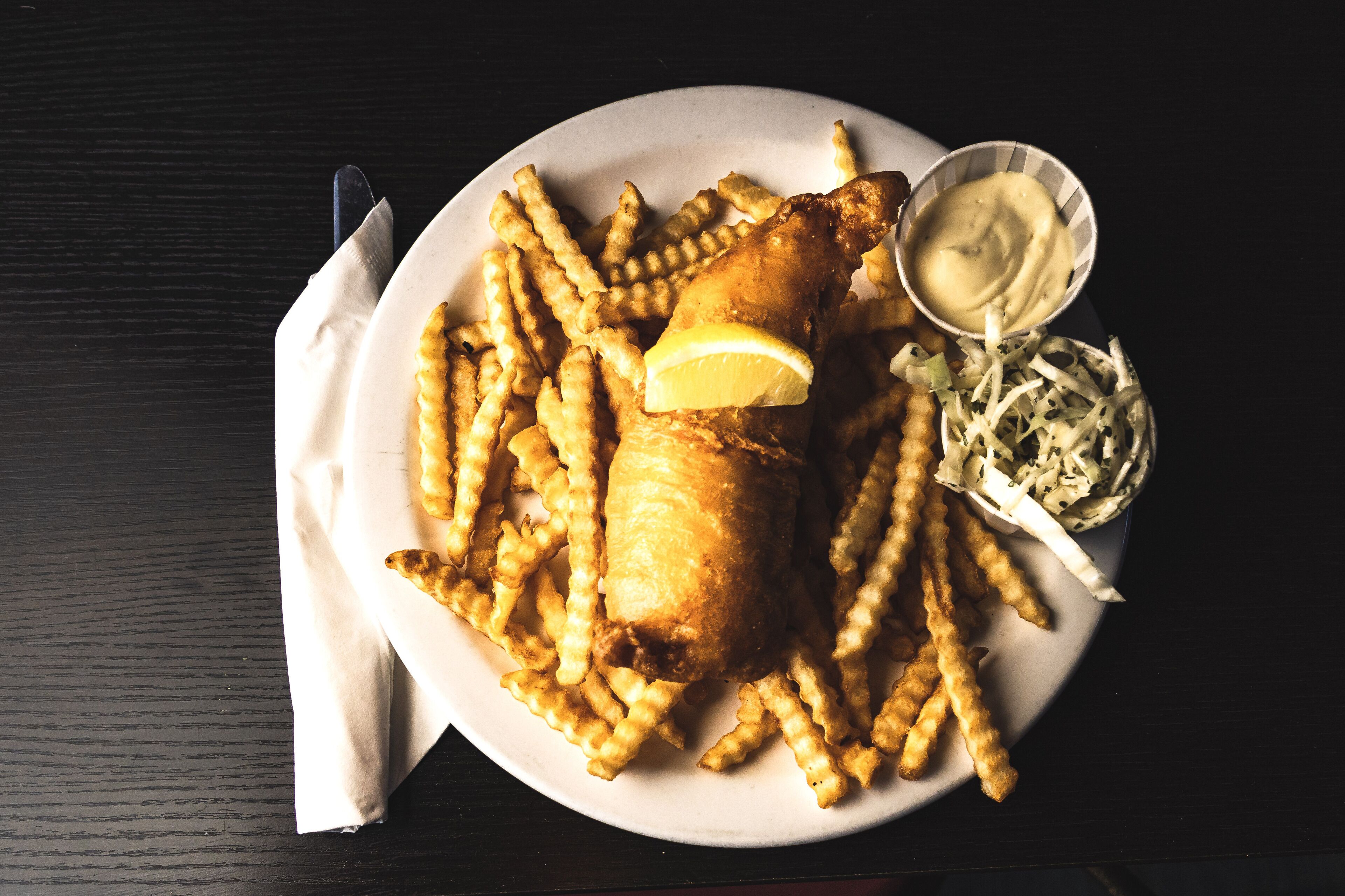 A plate of fish and chips with a lemon wedge, coleslaw, and a small bowl of sauce.