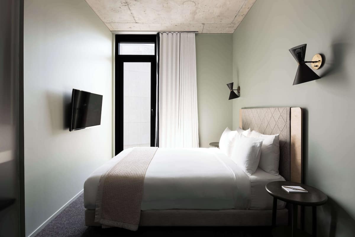 A hotel room with a bed, a bedside table, a TV mounted on the wall, and a window with curtains.