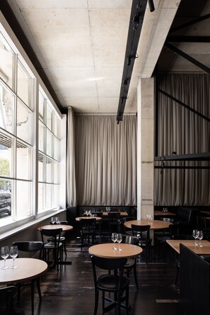 A restaurant with round tables and chairs, large windows with curtains, and a concrete ceiling.