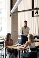 Interior de restaurante con un camarero sosteniendo una botella, tres comensales sentados en una mesa con platos y vasos.