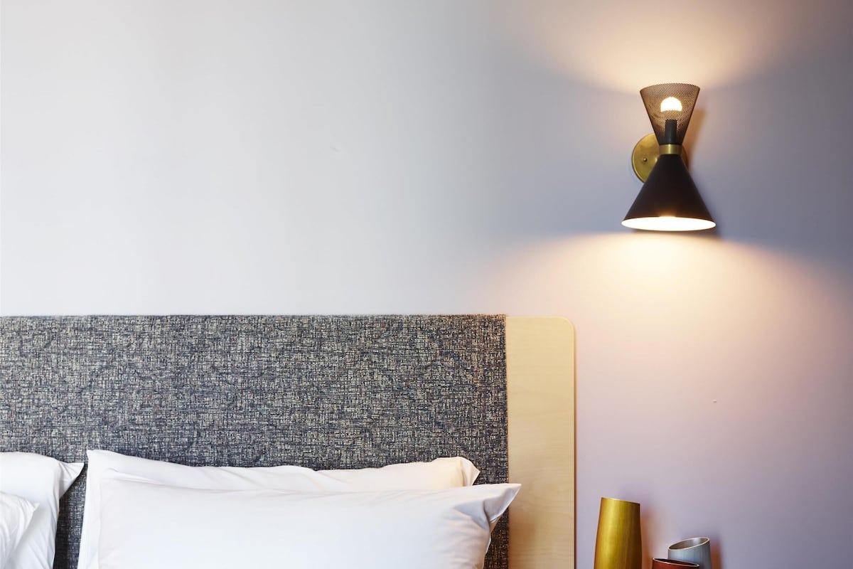 A neatly made bed with white pillows and a grey headboard, a small wooden side table with a lamp, and a magazine on the bed.
