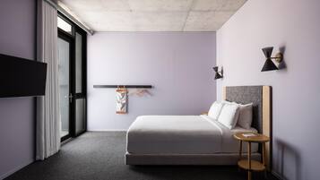 A modern hotel room with a bed, a small side table, a TV, and a window with curtains.