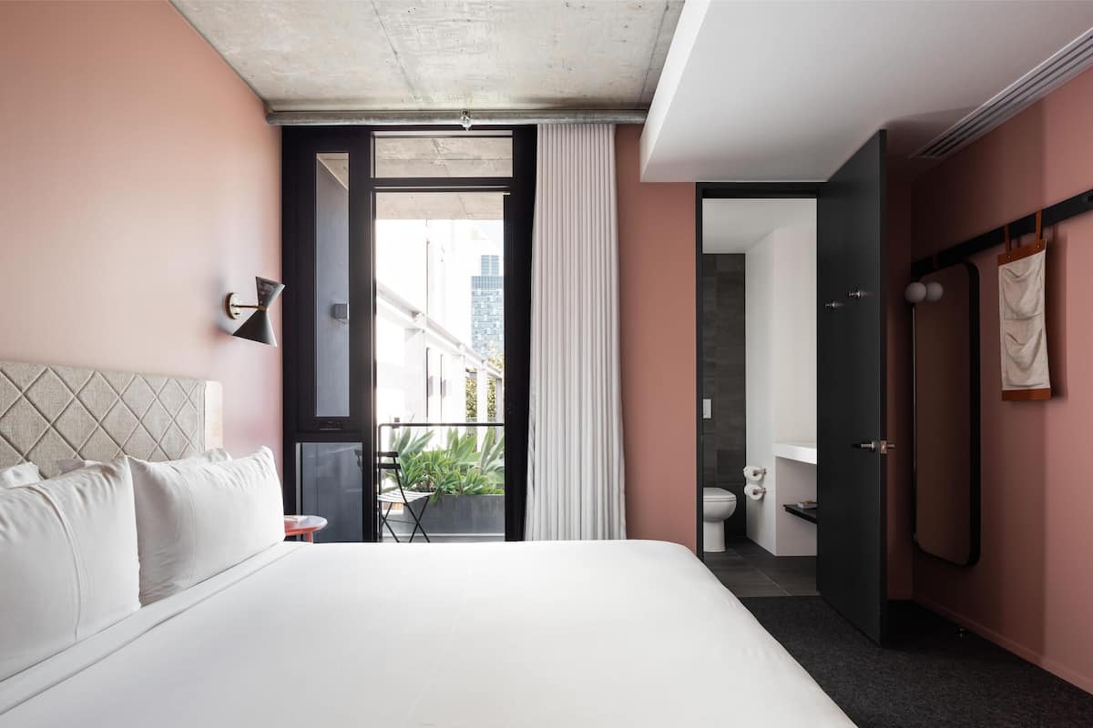 A modern hotel room with a large bed, a balcony with a view of buildings, a separate bathroom, and a wardrobe.