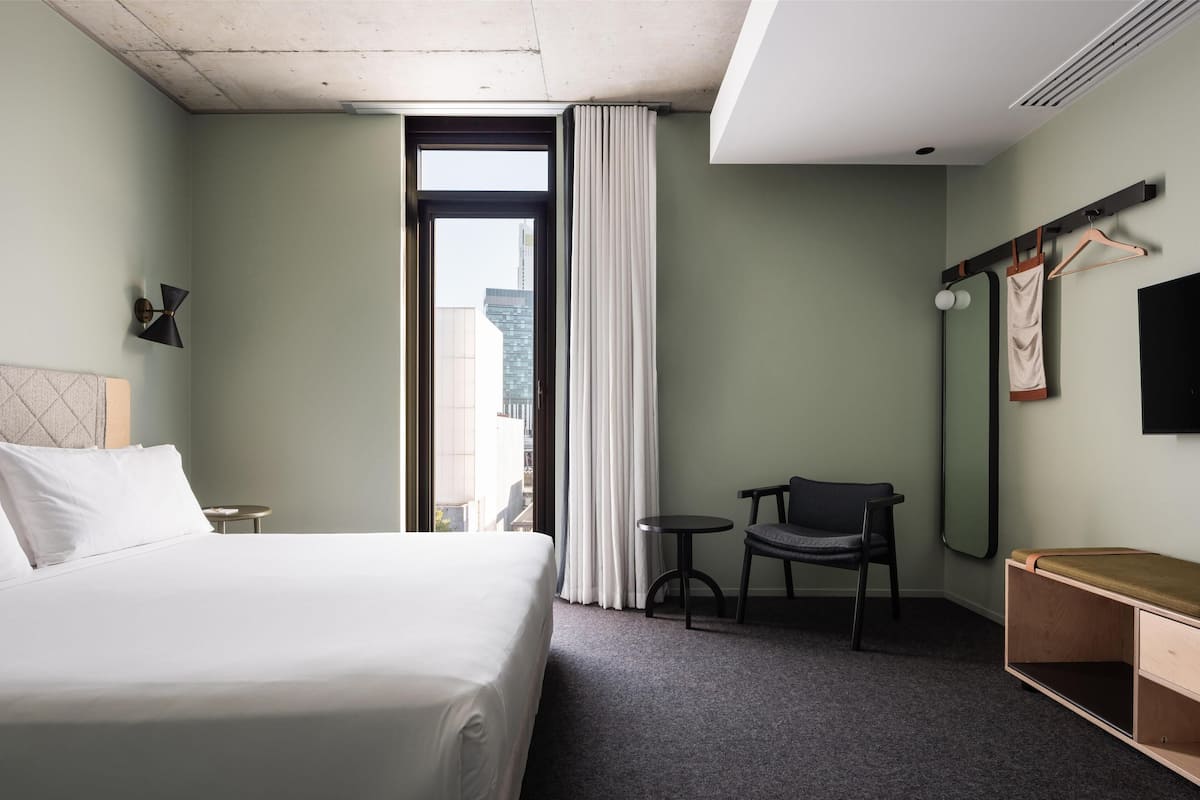 A hotel room with a bed, a small table, a chair, a desk, a mirror, and a window with curtains.