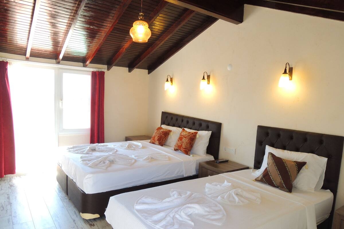 A hotel room with two beds, each with neatly folded towels, a wooden ceiling, and wall-mounted lights.