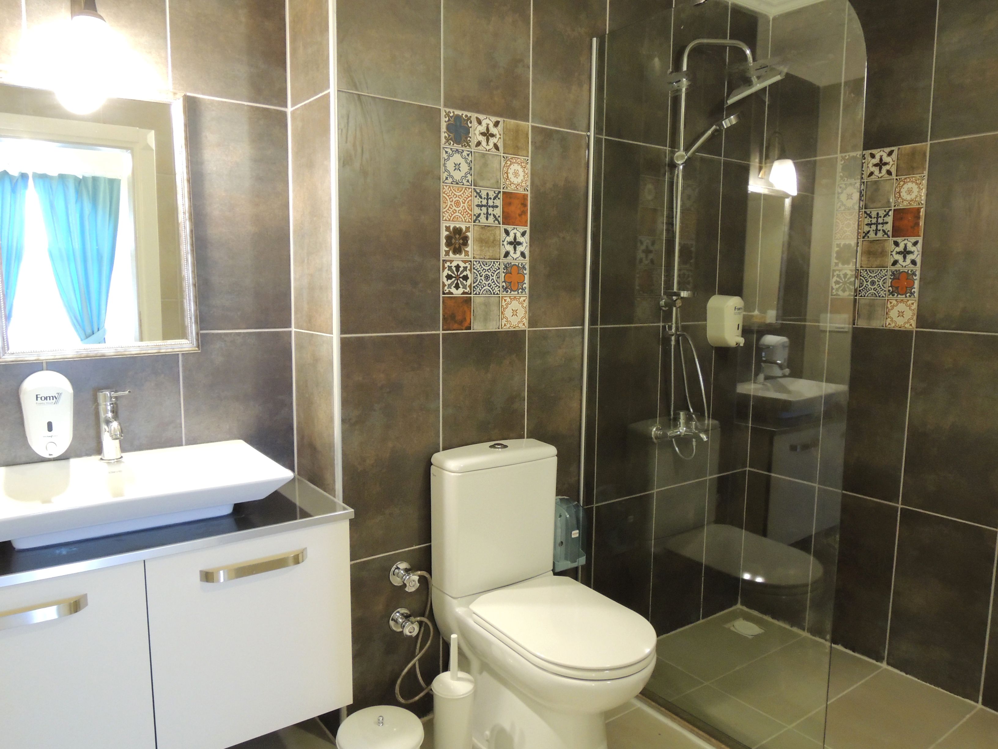 Une salle de bain avec un lavabo, des toilettes et une douche, le tout de couleur blanche. Les murs sont recouverts de carreaux aux motifs variés.