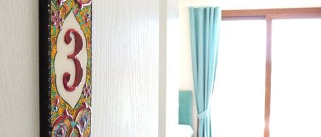 Une pièce avec une plaque numérotée fixée au mur, indiquant le numéro 3, une fenêtre avec des rideaux bleu clair et un luminaire suspendu.