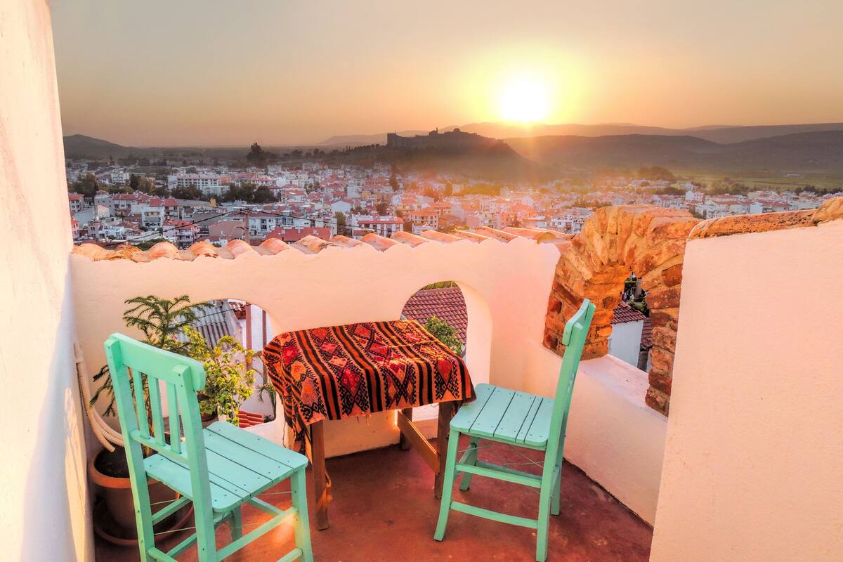 A balcony with a table and chairs, overlooking a cityscape during sunset.