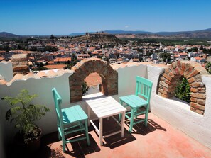 Eine Terrasse mit zwei türkisfarbenen Stühlen und einem Tisch, mit Blick auf eine Stadt mit historischen Gebäuden und einer Hügelkette in der Ferne.