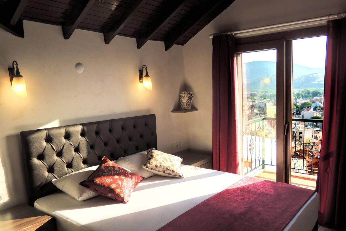 A bedroom with a tufted headboard, a bed with pillows, a nightstand, a wall-mounted light, a decorative item, and a balcony with a view.