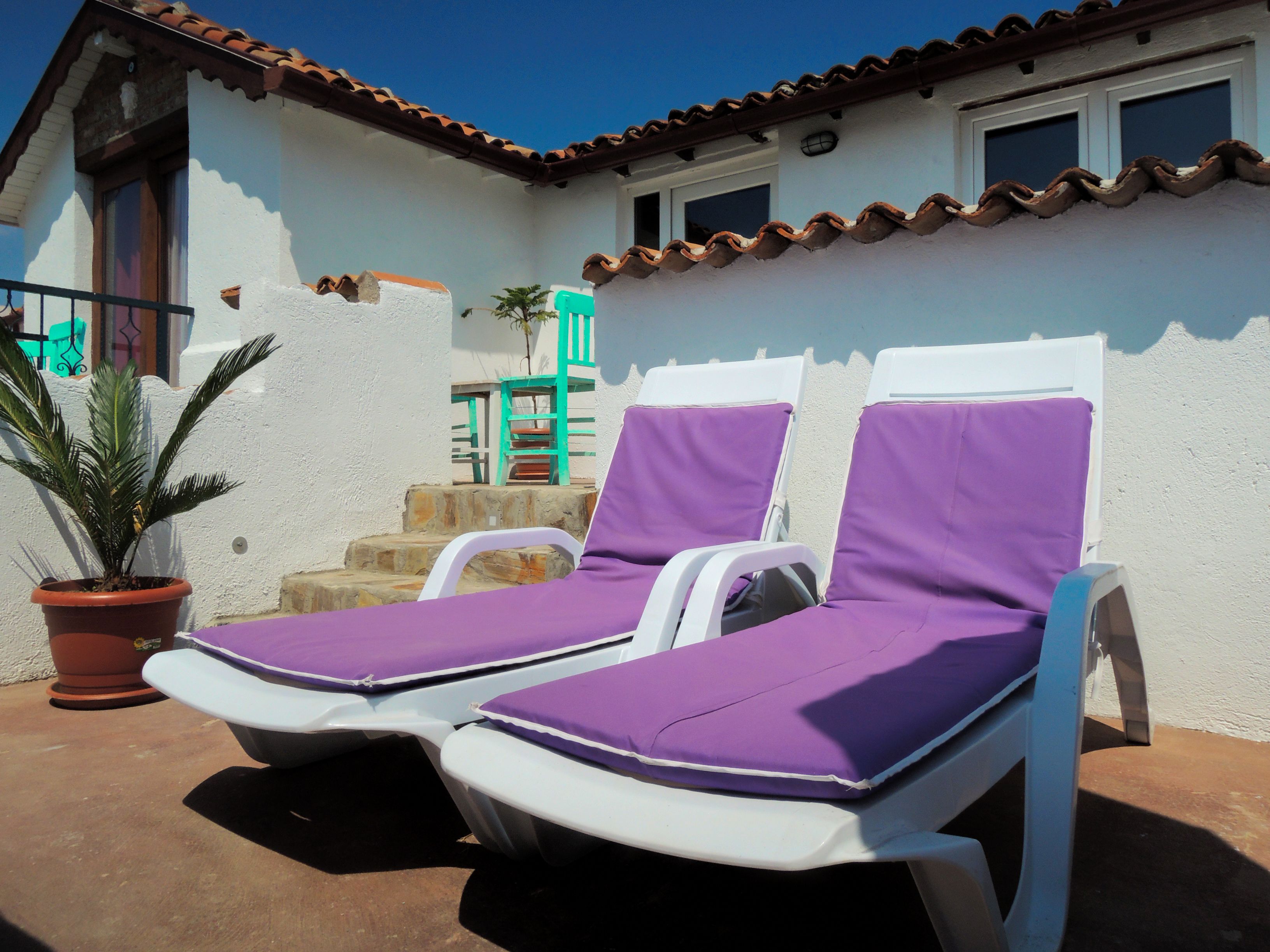 Two sun loungers with purple cushions outside a building with a tiled roof and white walls.