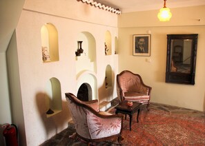 A room with a patterned carpet, two antique chairs, a fireplace, and a framed portrait on the wall.