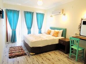 A bedroom with a bed, a desk, and a chair. There are curtains on the windows, an air conditioner, and a wall-mounted light fixture.