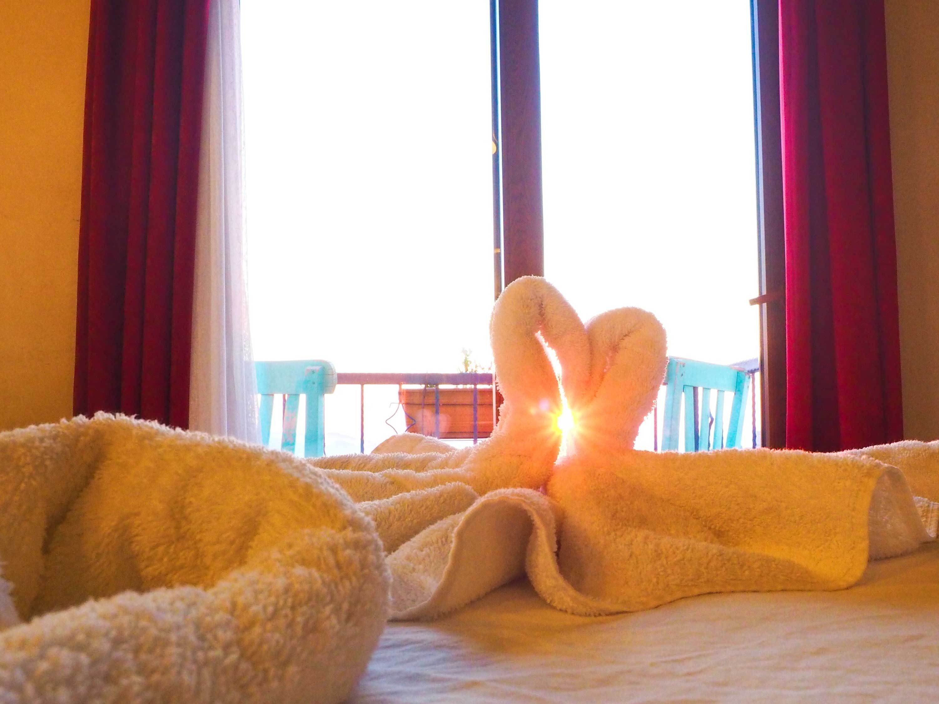 A bed with a heart-shaped towel arrangement and a view of a balcony with red curtains.