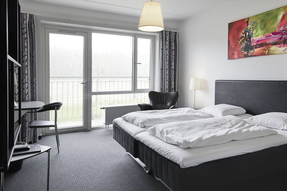 A hotel room with a bed, a desk, a chair, a radiator, and a large window.