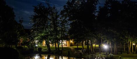 Une scène nocturne dans un parc, avec un étang, des arbres et un bâtiment illuminé.
