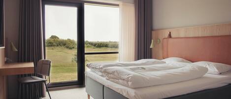 A hotel room with a bed, a desk, a chair, and a large window with a view of greenery.