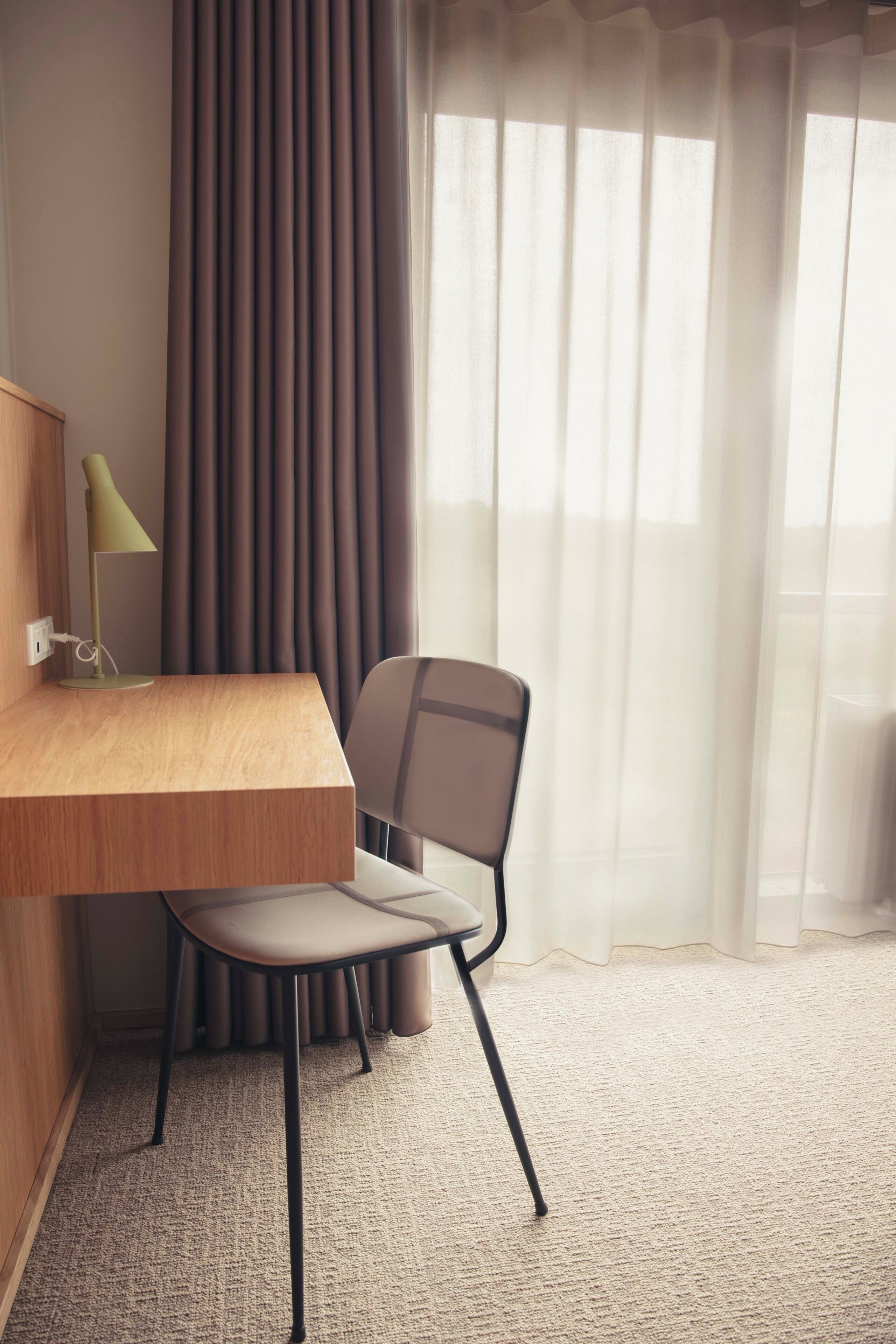 A hotel room with a wooden desk, a chair, a lamp, and a view through sheer curtains.