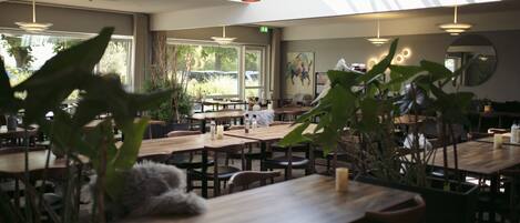 Un restaurante con mesas y sillas de madera, ventanas amplias y lámparas colgantes.