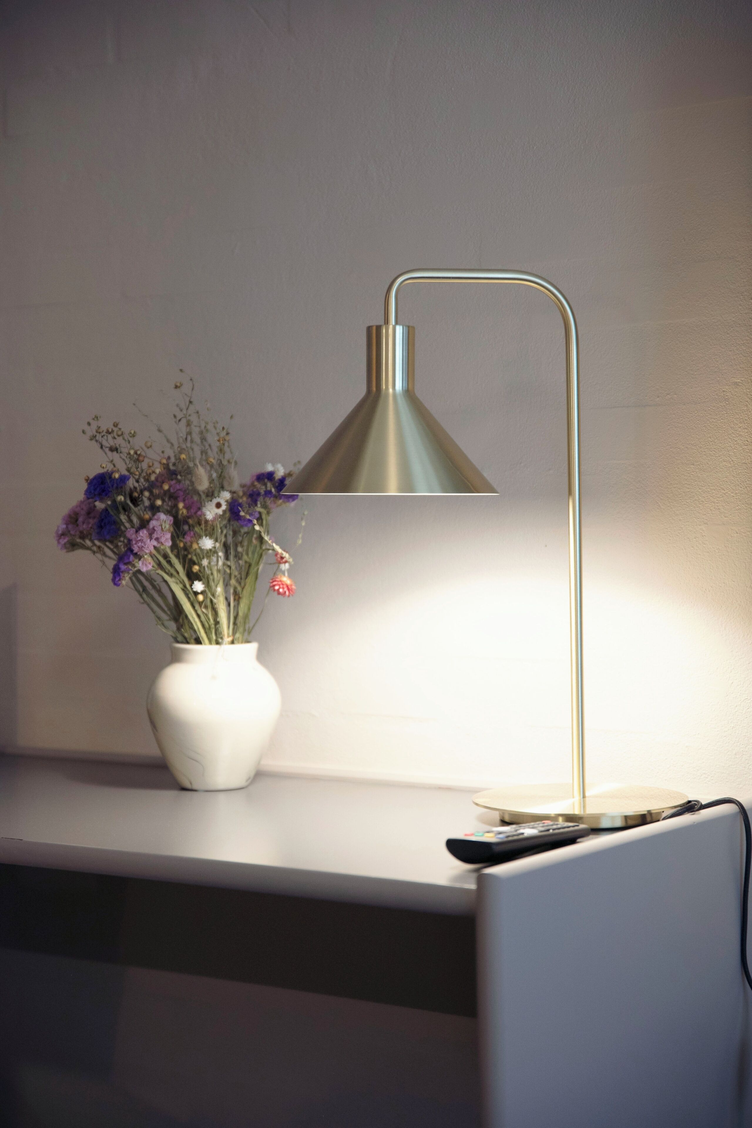 A desk with a vase of flowers, a lamp, and a remote control.