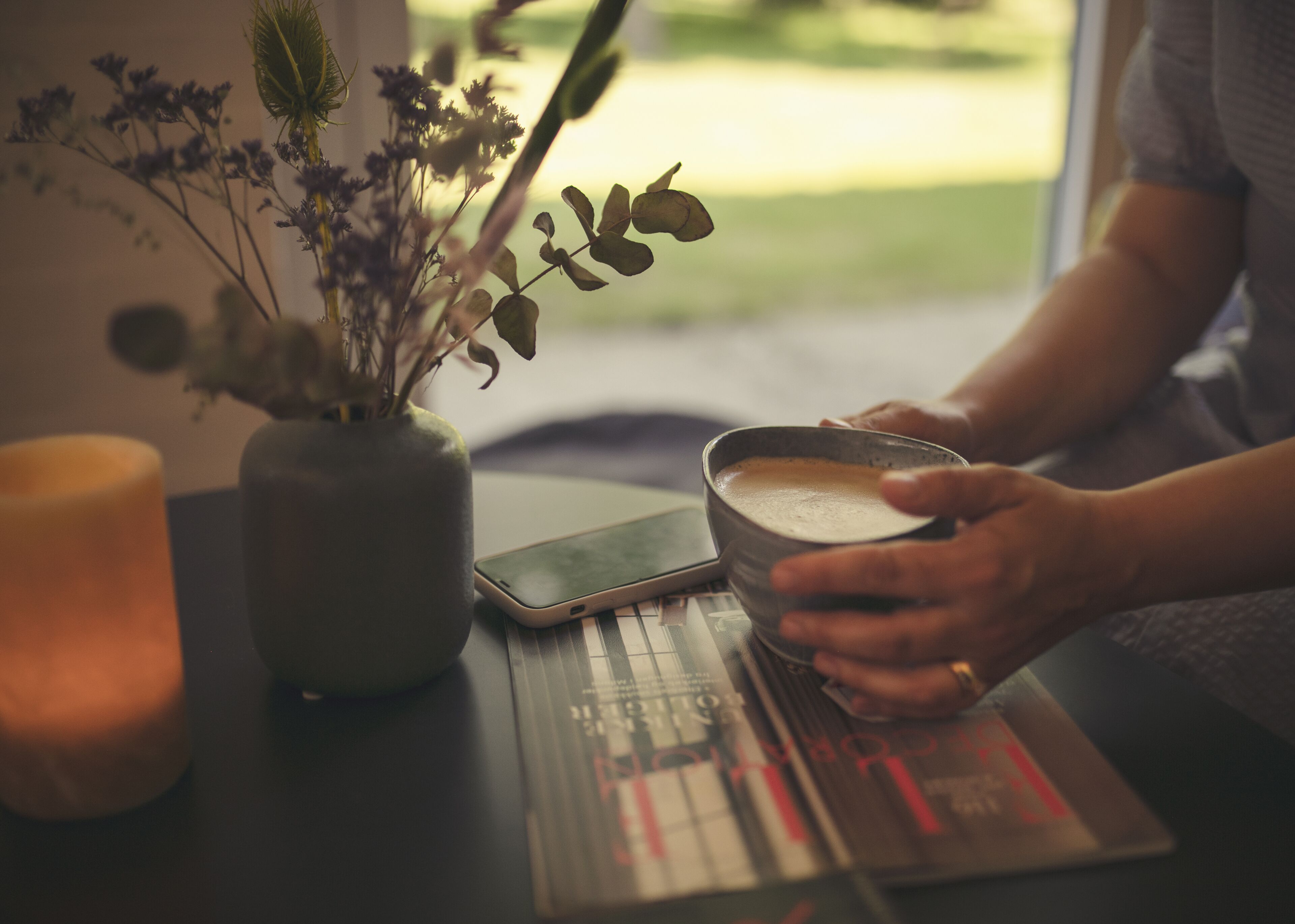 A person holding a cup of coffee, a smartphone, and a book on a table with a candle and a vase of flowers.