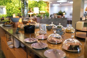 Montaje de buffet con diversos platos y condimentos, una estación de café y una pared verde de fondo.